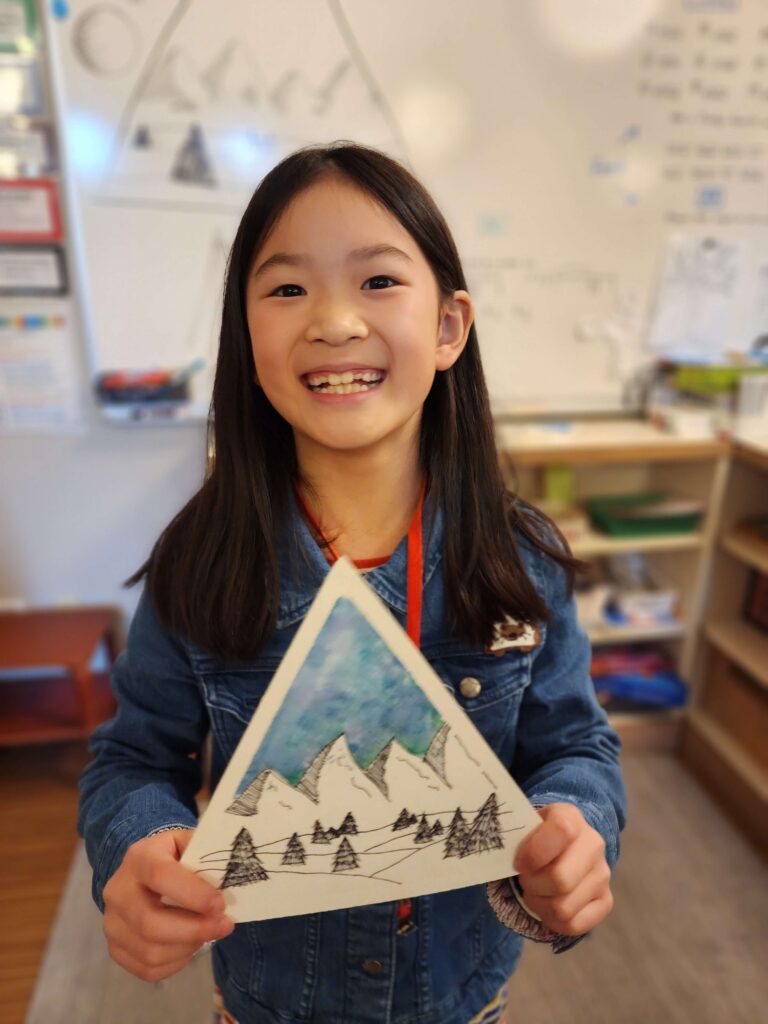 An elementary student proudly smiles while she shows off her watercolor painting of mountains.