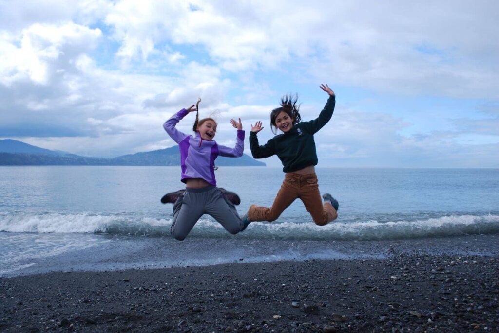 Two girls jump up together in the air by the ocean.