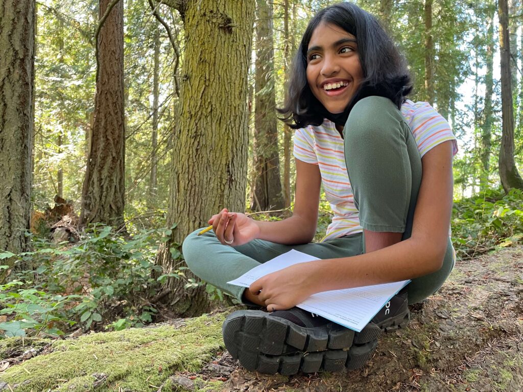A student sits on a tree log while writing in a worksheet.