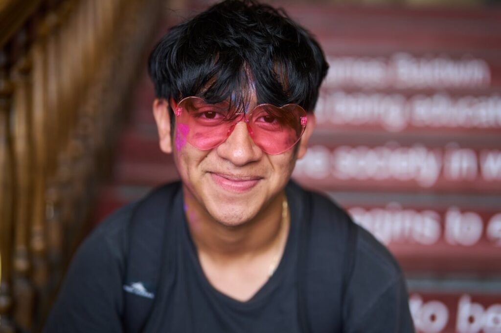 A high school boy smiles while wearing pink, heart shaped sunglasses.
