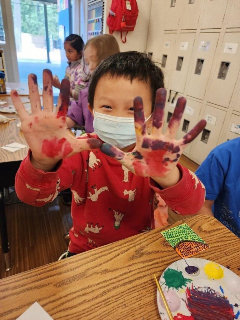 An elementary student smiles as he shows off his paint covered hands.