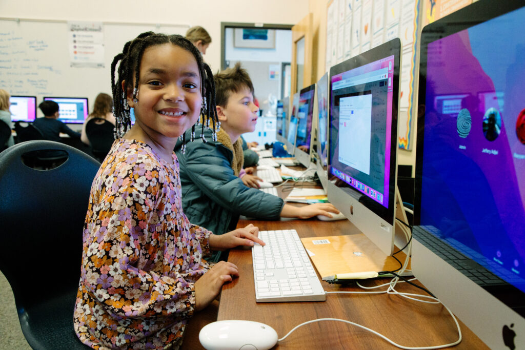 An elementary student typing on a computer smiles at the camera.